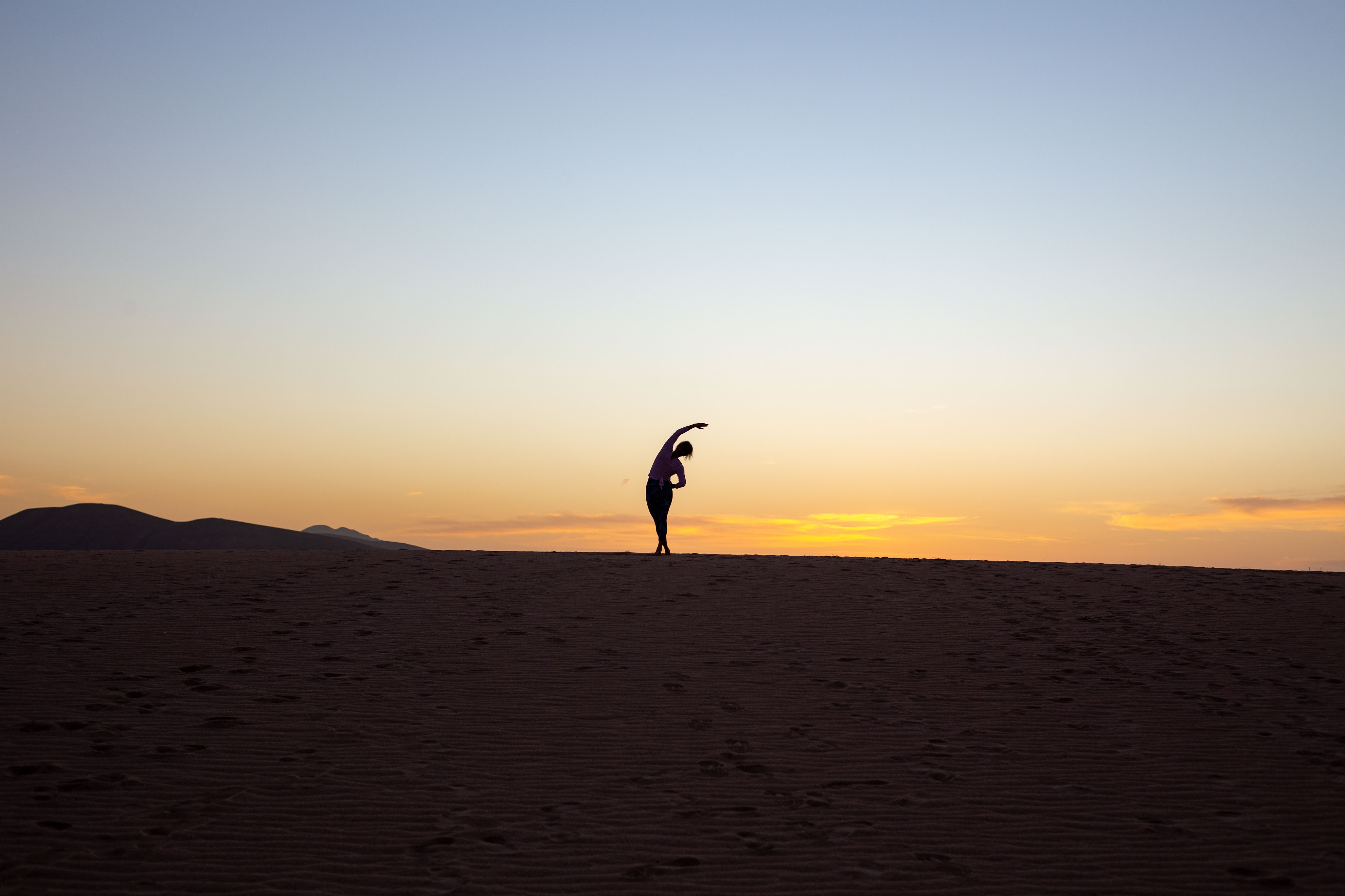 Eszter in a standing side bend on a dune at sunset, in silhouette against the sky
